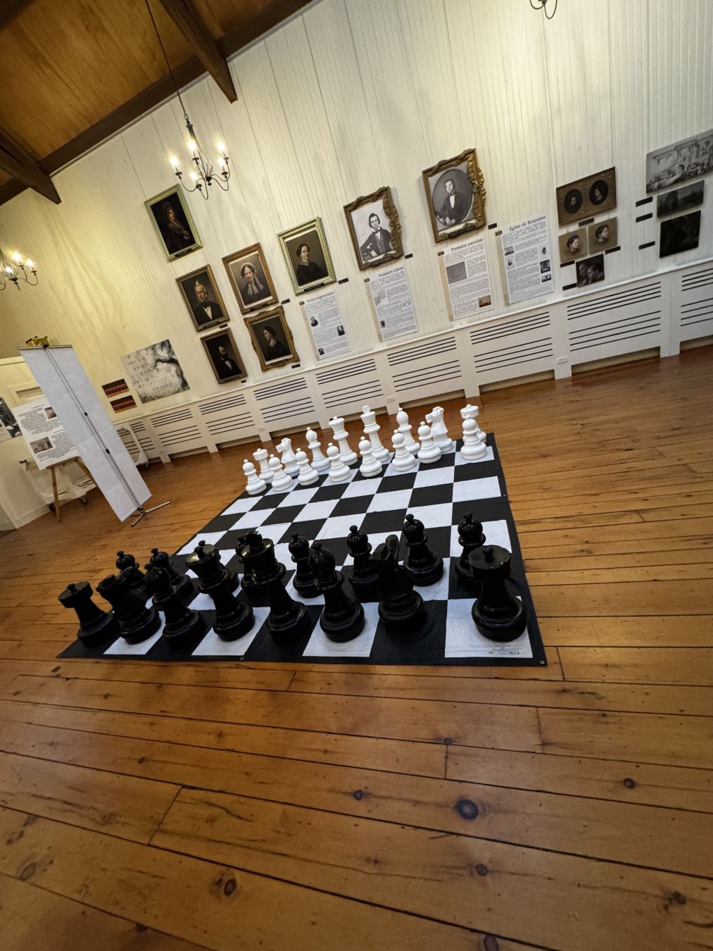 Giant chess board on a wooden floor inside the family museum. There are pictures of the Papineau family members on the wall.

The chess pieces are in their starting positions.
- - -
Un échiquier géant sur un plancher en bois à l'intérieur du musée familial. Il y a des photos des membres de la famille Papineau sur le mur.

Les pièces d'échecs sont dans leurs positions de départ.