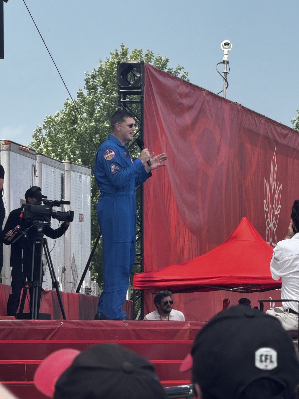 Colonel Jeremy Hansen speaking on stage. He's wearing his blue spacesuit, with Canadian space mission badges.

There are red Canada Day backdrops behind him
- - -
Le colonel Jeremy Hansen parle sur scène. Il porte sa combinaison spatiale bleue, avec les insignes de la mission spatiale canadienne.

Il y a des toiles de fond rouges de la Fête du Canada derrière lui